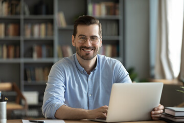 Good-looking millennial office employee in glasses sitting at desk in front of laptop smiling...