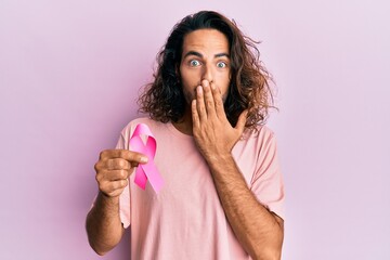 Young handsome man with long hair holding pink cancer ribbon covering mouth with hand, shocked and afraid for mistake. surprised expression