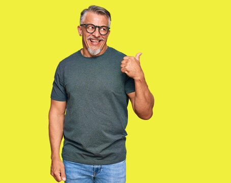 Middle age grey-haired man wearing casual clothes and glasses smiling with happy face looking and pointing to the side with thumb up.
