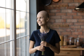 Life goes on. Happy young woman recovering from cancer standing at kitchen holding lavender bouquet...