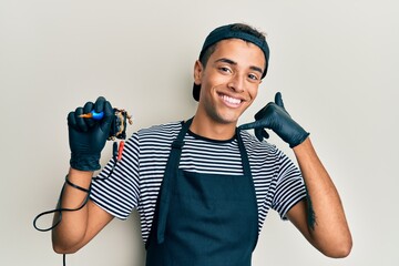 Young handsome african american man tattoo artist wearing professional uniform and gloves holding tattooer machine smiling doing phone gesture with hand and fingers like talking on the telephone. 