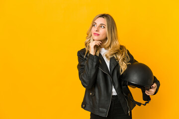 Young blonde caucasian biker woman holding helmet looking sideways with doubtful and skeptical expression.