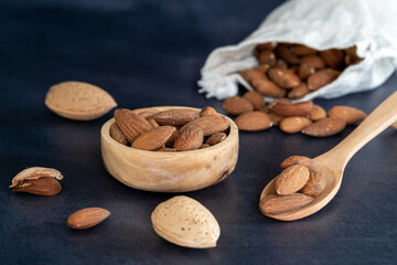 Almonds in a wooden bowl and on a wooden spoon on a black wooden table. some fruits are in a cotton mesh eco bag