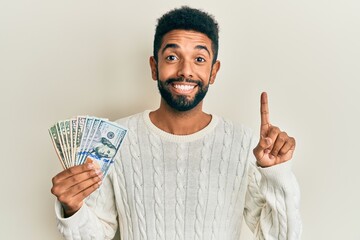 Handsome young hispanic man holding dollars smiling happy pointing with hand and finger to the side