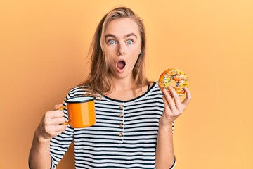 Beautiful blonde woman eating doughnut and drinking coffee afraid and shocked with surprise and amazed expression, fear and excited face.