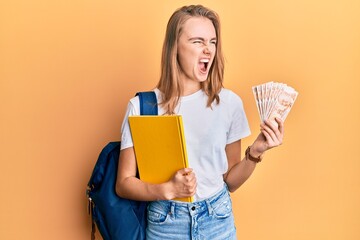 Beautiful blonde woman wearing student backpack and holding 50 turkish lira angry and mad screaming frustrated and furious, shouting with anger. rage and aggressive concept.