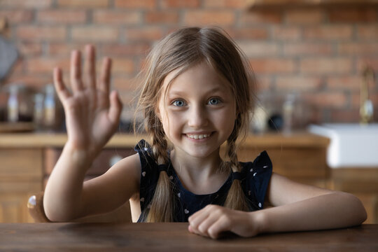 Hello, that is me. Portrait of smiling little girl sitting at table at home looking at camera waving hand in greeting gesture, cute vlogger child recording new content on webcam welcoming subscribers