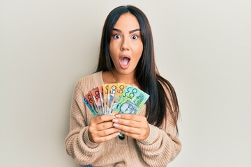Young beautiful hispanic girl holding australian dollars afraid and shocked with surprise and...
