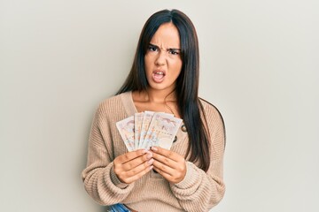 Young beautiful hispanic girl holding 10 united kingdom pounds banknotes in shock face, looking skeptical and sarcastic, surprised with open mouth