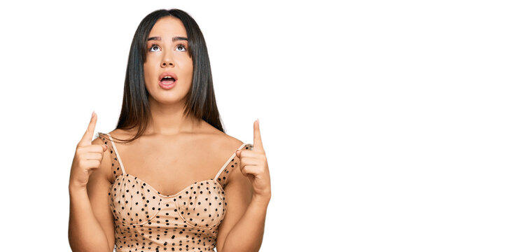 Young beautiful hispanic girl wearing sexy party dress amazed and surprised looking up and pointing with fingers and raised arms.