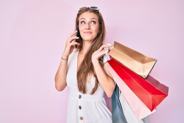 Young blonde girl holding shopping bags talking on the smartphone smiling looking to the side and staring away thinking.