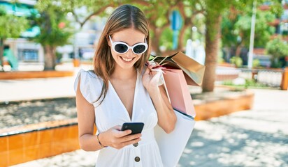 Young blonde woman smiling happy holding shopping bags and using smartphone at street of city