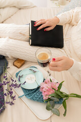 Woman dressed on warm loungewear on bed with a closed book over her lap. She is holding a cup of foamy coffee.