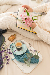 Woman reading a book on bed next to her coffee breakfast. She is holding a pretty bouquet of flowers with her left hand.