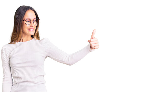 Beautiful brunette young woman wearing casual white sweater and glasses looking proud, smiling doing thumbs up gesture to the side