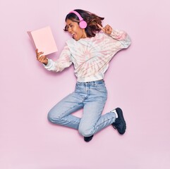 Naklejka premium Young beautiful latin woman listening to music using headphones smiling happy. Jumping with smile on face reading book over isolated pink background