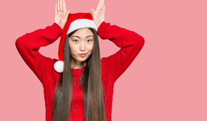 Young chinese woman wearing christmas hat doing bunny ears gesture with hands palms looking cynical...