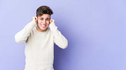 Young caucasian man isolated on purple background covering ears with hands.