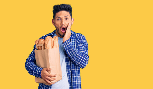 Handsome latin american young man holding paper bag with bread afraid and shocked, surprise and amazed expression with hands on face