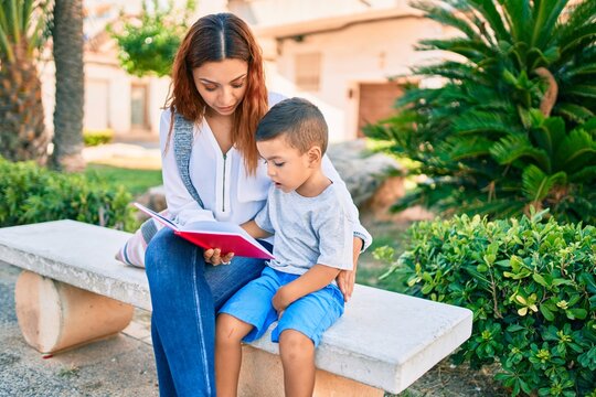 Adorable latin mother and son sitting on the bench and reading book at the park.