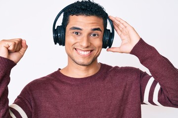 Young handsome hispanic man listening to music using headphones screaming proud, celebrating victory and success very excited with raised arm