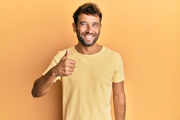 Handsome man with beard wearing casual yellow tshirt over yellow background doing happy thumbs up gesture with hand. approving expression looking at the camera showing success.