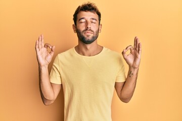 Handsome man with beard wearing casual yellow tshirt over yellow background relax and smiling with eyes closed doing meditation gesture with fingers. yoga concept.
