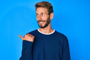 Handsome caucasian man with beard wearing casual sweater smiling with happy face looking and pointing to the side with thumb up.