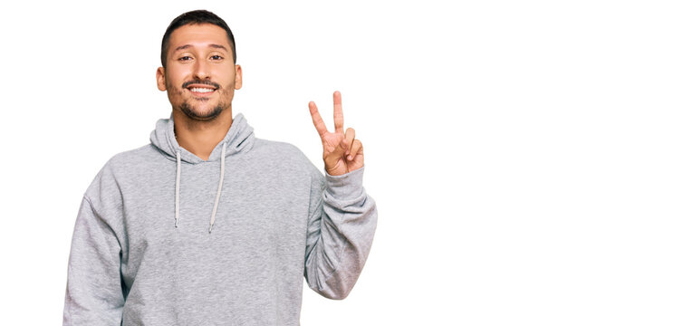 Handsome man with tattoos wearing casual sweatshirt smiling with happy face winking at the camera doing victory sign. number two.