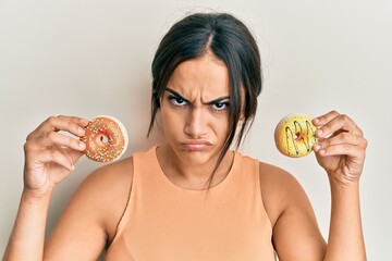 Young brunette woman holding tasty colorful doughnuts skeptic and nervous, frowning upset because...