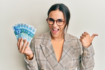 Young brunette woman holding hong kong 20 dollars banknotes pointing thumb up to the side smiling happy with open mouth