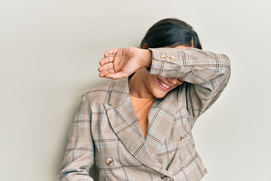 Young brunette woman wearing business jacket and glasses covering eyes with arm smiling cheerful and funny. blind concept.