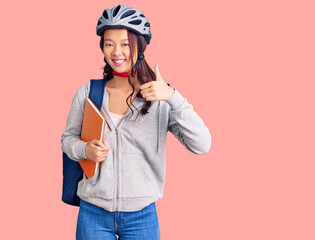 Young beautiful chinese girl wearing student backpack and bike helmet holding book smiling happy and positive, thumb up doing excellent and approval sign