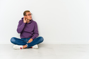 Young caucasian man sitting on the floor trying to listening a gossip.