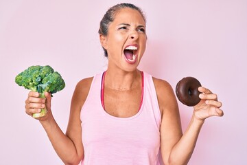 Middle age hispanic woman holding broccoli and chocolate donut angry and mad screaming frustrated...