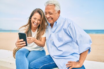 Middle age hispanic couple using smartphone sitting on the bench at the beach.