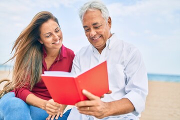 Middle age hispanic couple reading book sitting on the bench at the beach.