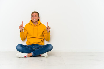 Young caucasian man sitting on the floor indicates with both fore fingers up showing a blank space.