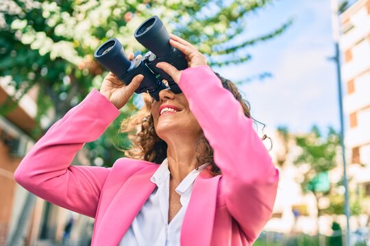 Middle age hispanic businesswoman smiling happy looking for new opportunity using binoculars at the city.