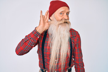 Old senior man with grey hair and long beard wearing hipster look with wool cap smiling with hand over ear listening an hearing to rumor or gossip. deafness concept.