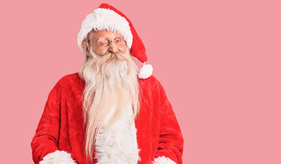 Old senior man with grey hair and long beard wearing traditional santa claus costume winking...