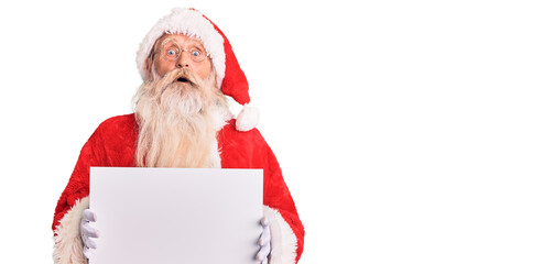 Old senior man with grey hair and long beard wearing santa claus costume holding banner scared and amazed with open mouth for surprise, disbelief face