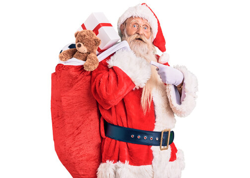 Old senior man with grey hair and long beard wearing santa claus costume holding bag with presents smiling happy pointing with hand and finger