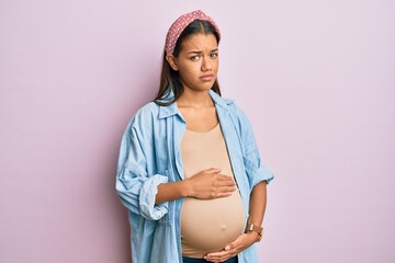 Beautiful hispanic woman expecting a baby, touching pregnant belly clueless and confused expression. doubt concept.