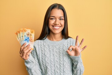 Beautiful hispanic woman holding 10 swiss franc banknotes doing ok sign with fingers, smiling friendly gesturing excellent symbol