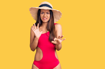 Young beautiful latin girl wearing swimwear and summer hat disgusted expression, displeased and fearful doing disgust face because aversion reaction. with hands raised