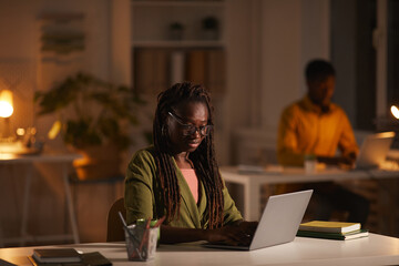 Portrait of contemporary African-American woman using laptop while working late in dark office,...