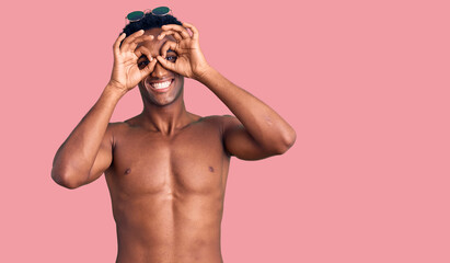 African handsome man wearing swimsuit and sunglasses doing ok gesture like binoculars sticking tongue out, eyes looking through fingers. crazy expression.