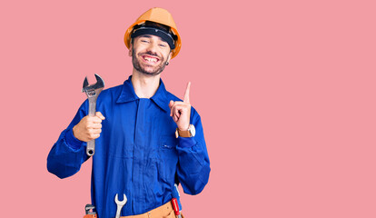 Young hispanic man wearing electrician uniform holding wrench surprised with an idea or question pointing finger with happy face, number one