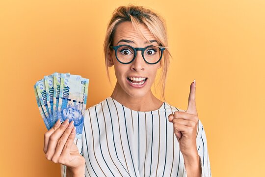 Beautiful blonde woman holding south african 100 rand banknotes smiling with an idea or question pointing finger with happy face, number one